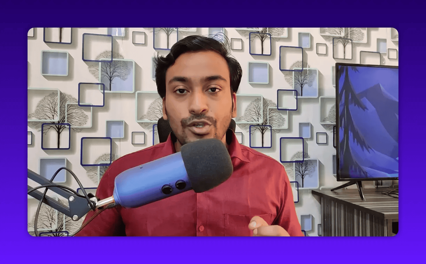 Host speaking into a blue microphone with a patterned wall background and a monitor on the right